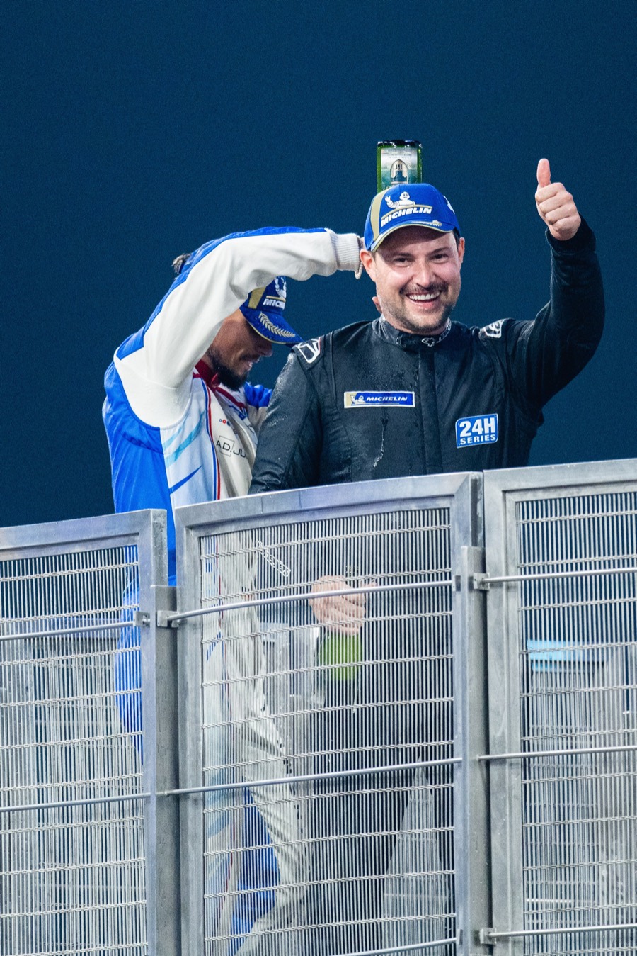 Champagne on the podium at night, a thumbs-up over the rail