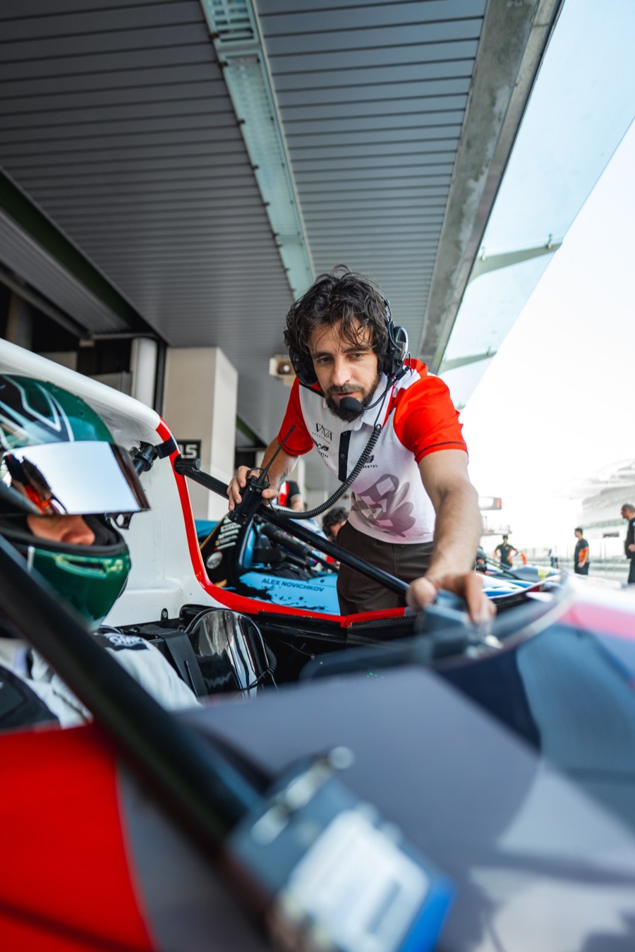 A mechanic reaching deep into the cockpit before a session, the driver's helmet on the seat beside him