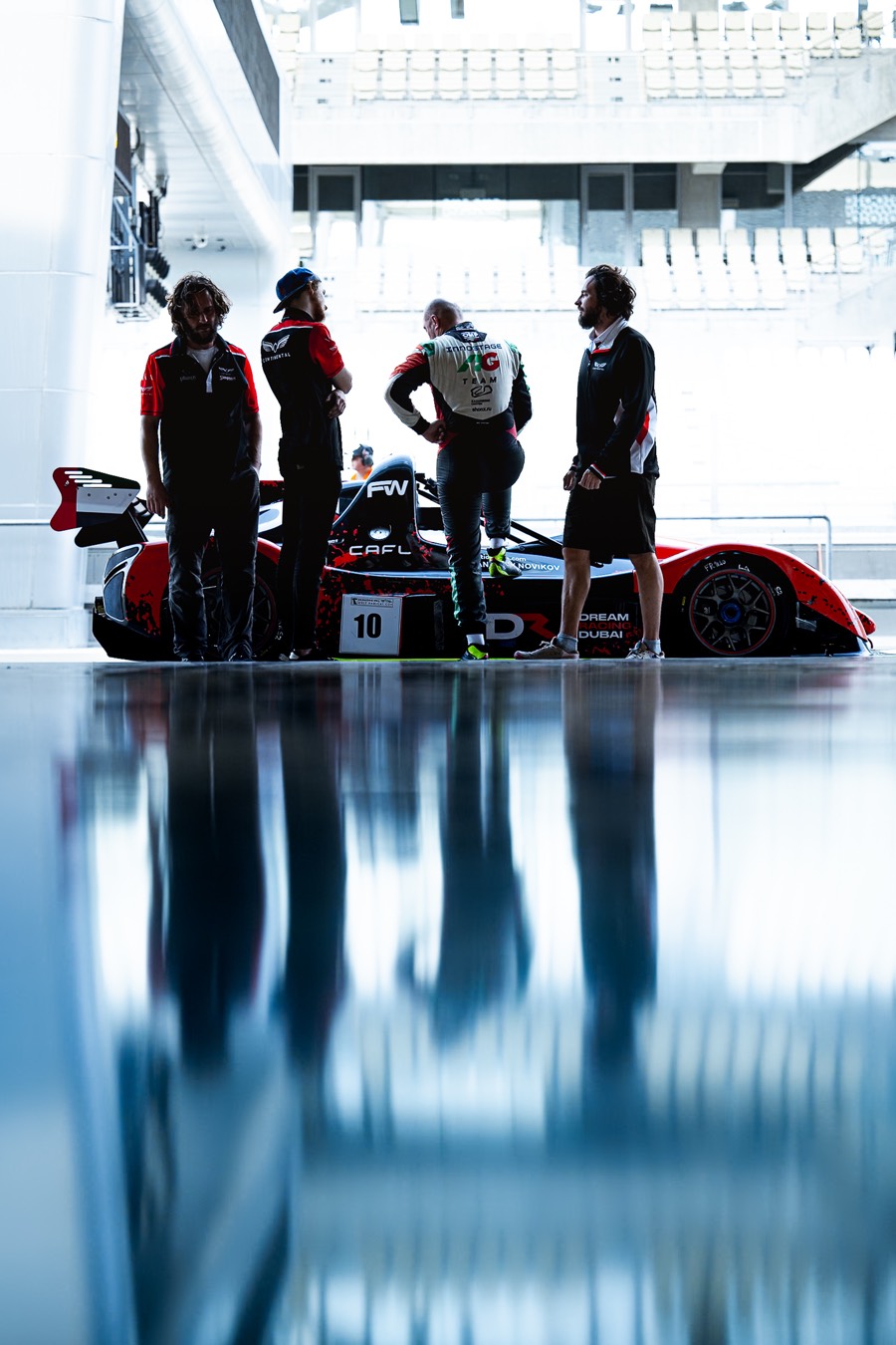 Four of the crew standing round the prototype in the garage, the whole scene doubled in the polished floor