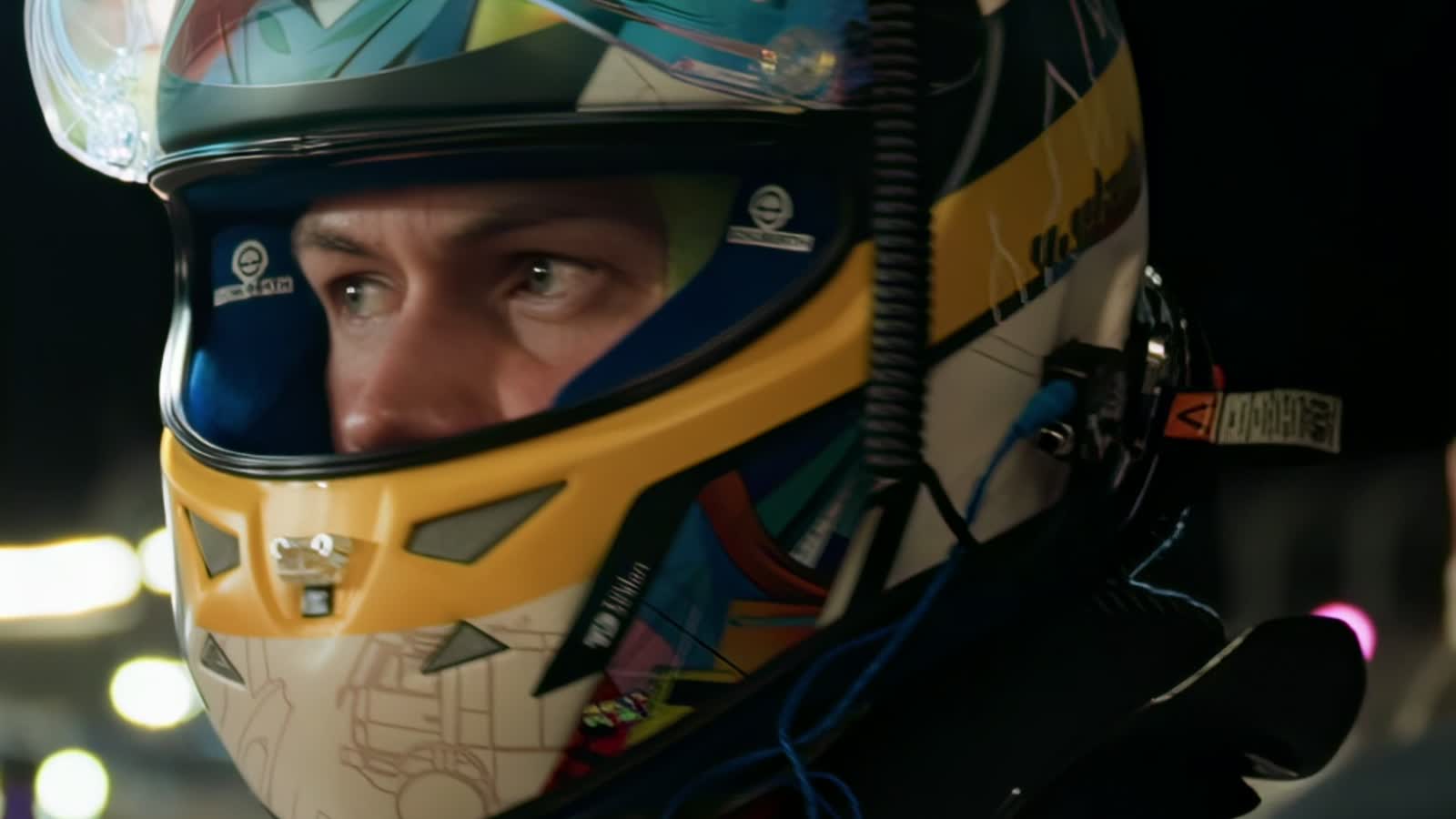 A driver in a helmet, close, under the pit-lane lights.